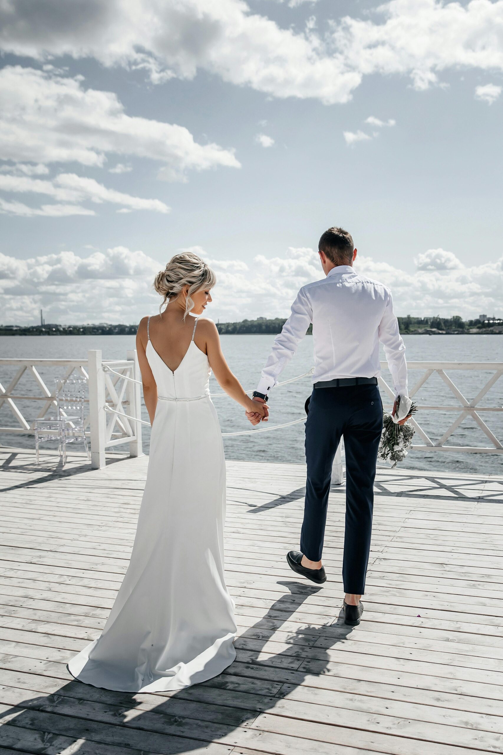 Bride and groom holding hands on a dock, with a serene water view on a sunny day.